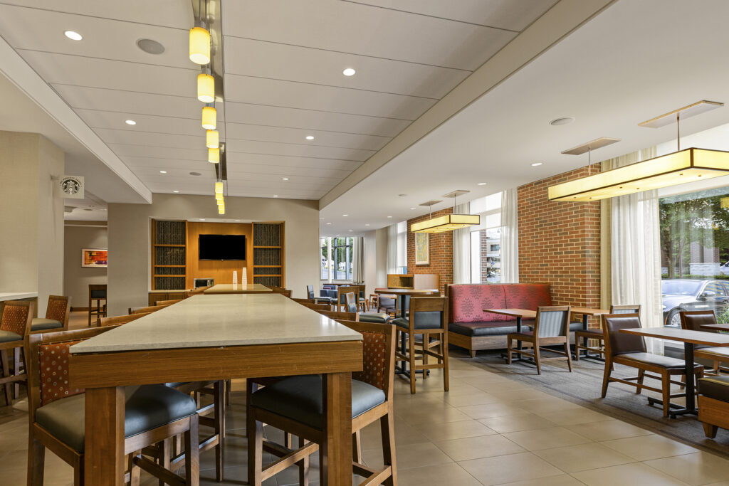 A modern hotel lounge at Hyatt Place in Bloomington, Illinois, features wooden furniture, cushioned chairs, and tall ceilings. Overhead lights illuminate the space, while large windows along a brick wall allow natural light in. Tables are arranged for group seating or individual use.