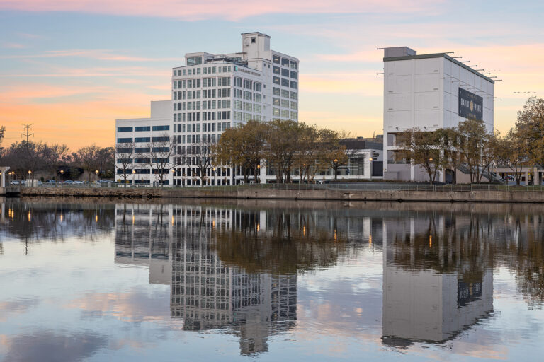 The reflection of modern buildings, including the Hilton, dances on a calm river at sunset. The sky is painted in soft pink and blue hues as trees line the riverbank, creating a tranquil scene mirrored perfectly in the water.