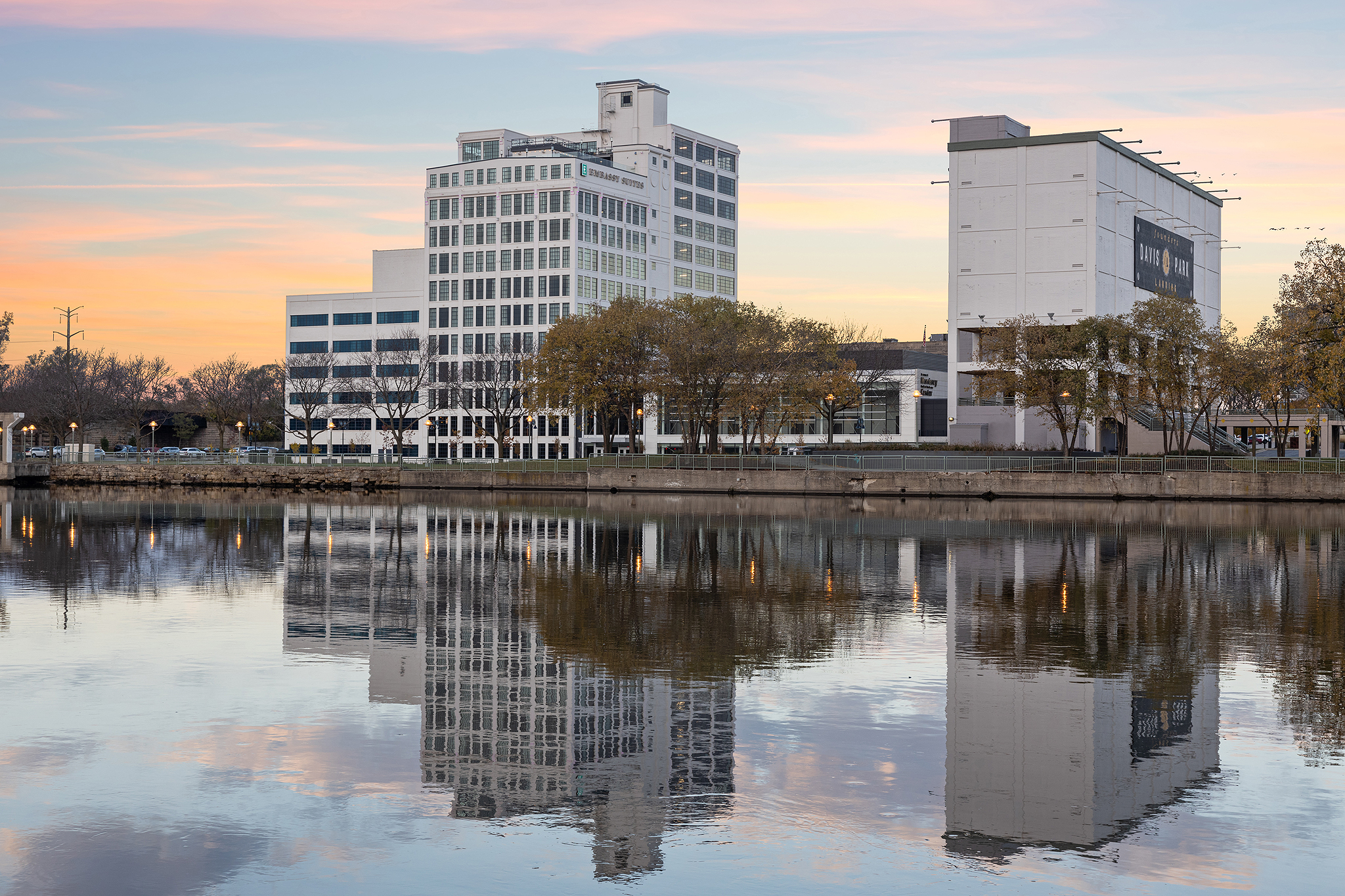 The reflection of modern buildings, including the Hilton, dances on a calm river at sunset. The sky is painted in soft pink and blue hues as trees line the riverbank, creating a tranquil scene mirrored perfectly in the water.