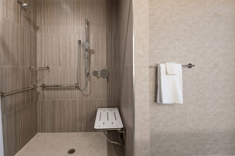 A modern shower at the Rockford Embassy Suites boasts beige tiles, a handheld showerhead on a vertical rail, and a foldable white bench. Multiple grab bars ensure safety, while a white towel hangs elegantly on a rod outside the shower area.