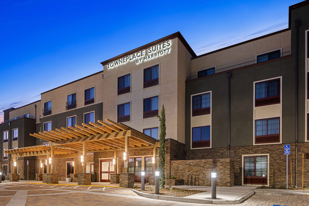 Exterior of a modern TownePlace Suites by Marriott hotel in San Luis Obispo, California, at dusk. The building showcases a sleek design with a pergola entrance and illuminated signage. The deep blue evening sky creates the perfect backdrop for this contemporary getaway.