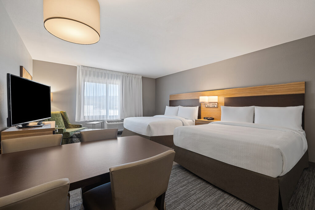 A modern TownePlace Suites hotel room in San Luis Obispo features two double beds, a desk with chairs, a flat-screen TV, and a window with sheer curtains. The room's neutral tones and clean lines create a comfortable and inviting atmosphere perfect for any Marriott guest.