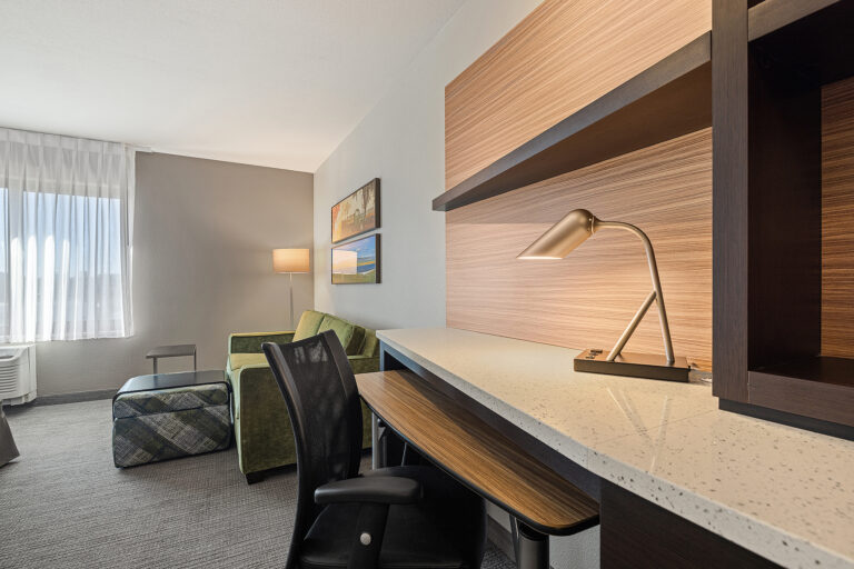 A modern Towneplace Suites hotel room in San Luis Obispo features a desk with a lamp and chair, a green sofa, an ottoman, and a floor lamp. Natural light streams through sheer curtains by the window, highlighting two framed artworks on the wall.