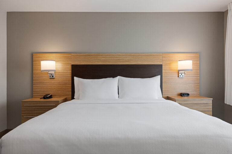 A modern Marriott hotel room in San Luis Obispo, California, with a large bed featuring white linens. The wooden headboard includes built-in nightstands on each side, each with a lamp and a phone on the left nightstand. The wall is painted in a neutral tone.