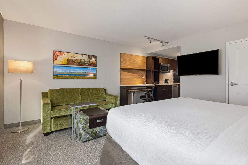 The modern Towneplace Suites hotel room in San Luis Obispo features a king-size bed, green sofa, and floor lamp. Wall art showcases a landscape. A small work area has a desk, chair, and shelves near the wall-mounted TV. In the corner is a kitchenette with a microwave and mini fridge.