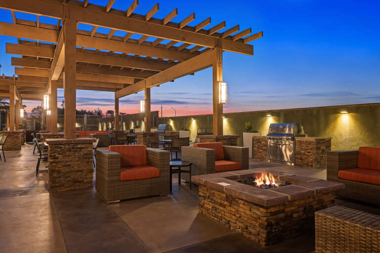 The outdoor patio at Towneplace Suites Marriott in California features a stone fire pit surrounded by wicker chairs with vibrant orange cushions. A wooden pergola stands overhead, while a grill awaits in the background, all beneath a dusk sky painted in shades of blue and orange.