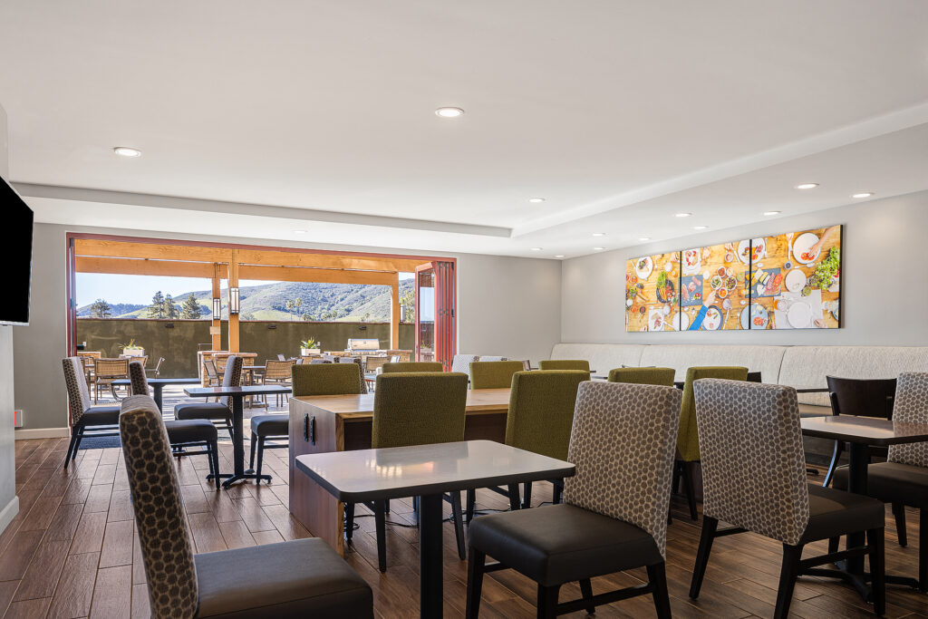 A modern dining area in TownePlace Suites, featuring wooden flooring, various patterned chairs, and several tables. A large, colorful painting decorates the wall. Glass doors lead to an outdoor patio with a view of the San Luis Obispo mountains under California's clear sky.