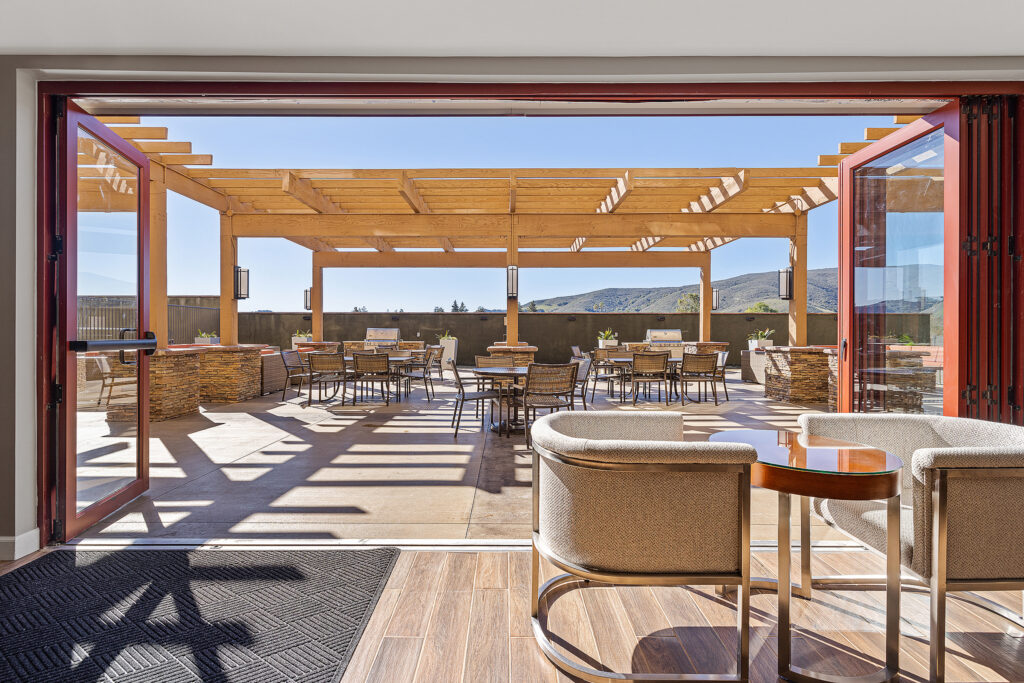 An open patio area at TownePlace Suites, San Luis Obispo features wooden tables and chairs under a wooden pergola. The bright blue sky and distant mountains enhance the view. The space is well-lit, with Marriott's signature indoor seating leading to the inviting patio.