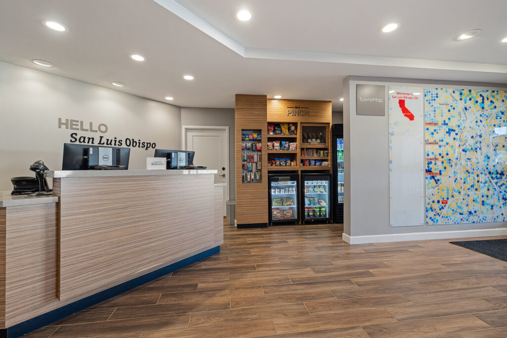 A reception area at TownePlace Suites features a wooden counter and a "Hello San Luis Obispo" sign. A snack and drink section complements the California map on the wall. Bright lighting and wooden flooring enhance the warm, welcoming Marriott atmosphere.