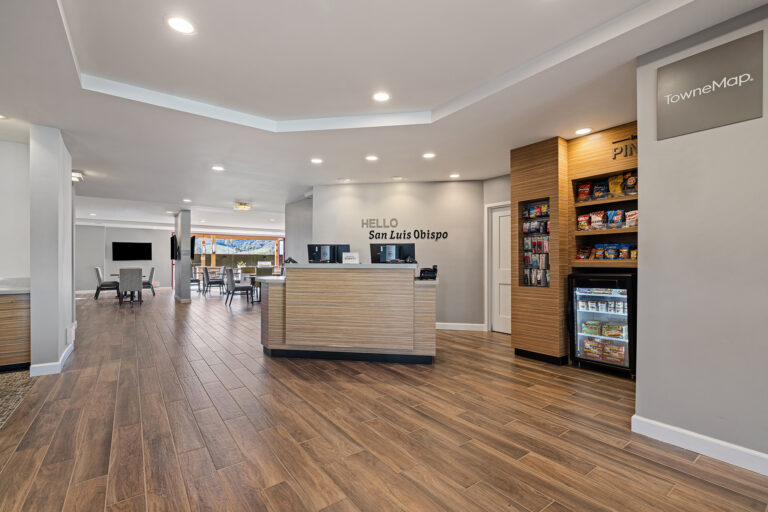 The TownePlace Suites by Marriott lobby in San Luis Obispo boasts modern decor, a sleek reception desk, and wood flooring. Visitors can enjoy a snack area with shelves and a fridge. "Hello San Luis Obispo" graces the wall behind the desk, while a cozy seating area invites relaxation in the background.