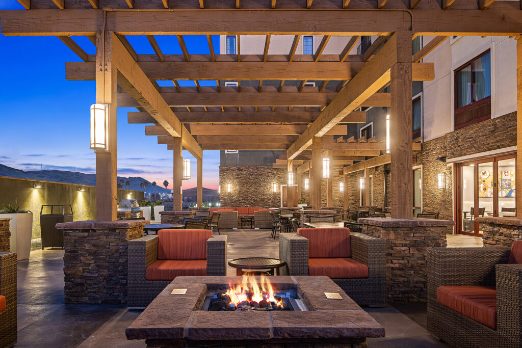 A cozy outdoor patio at twilight, nestled in California, features wicker furniture with red cushions around a lit fire pit. Stone pillars support wooden pergolas, and lantern-style lights create a warm ambiance. Hills are visible in the background, enhancing the serene Marriott atmosphere.