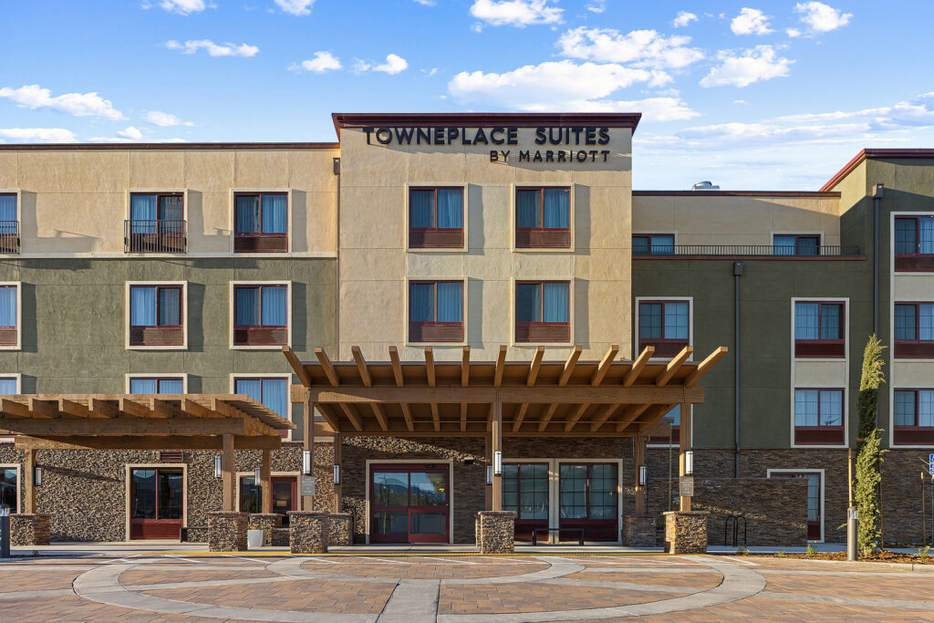 Exterior view of a TownePlace Suites by Marriott hotel in San Luis Obispo, California. The building features beige and green walls with multiple windows. A wooden pergola extends over the entrance, set against a clear sky with a few clouds.