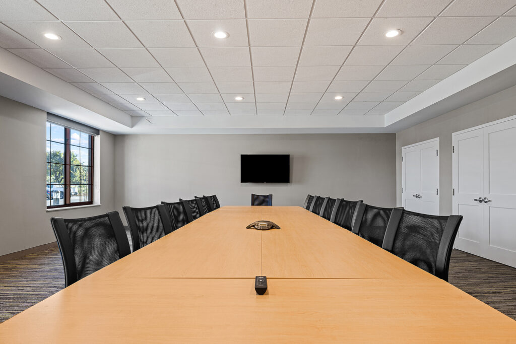 A modern conference room at Marriott in San Luis Obispo features a long wooden table surrounded by black mesh chairs. The room boasts a flat-screen TV on a gray wall and large windows that flood the space with California sunshine. Recessed lighting and acoustic tiles grace the ceiling.
