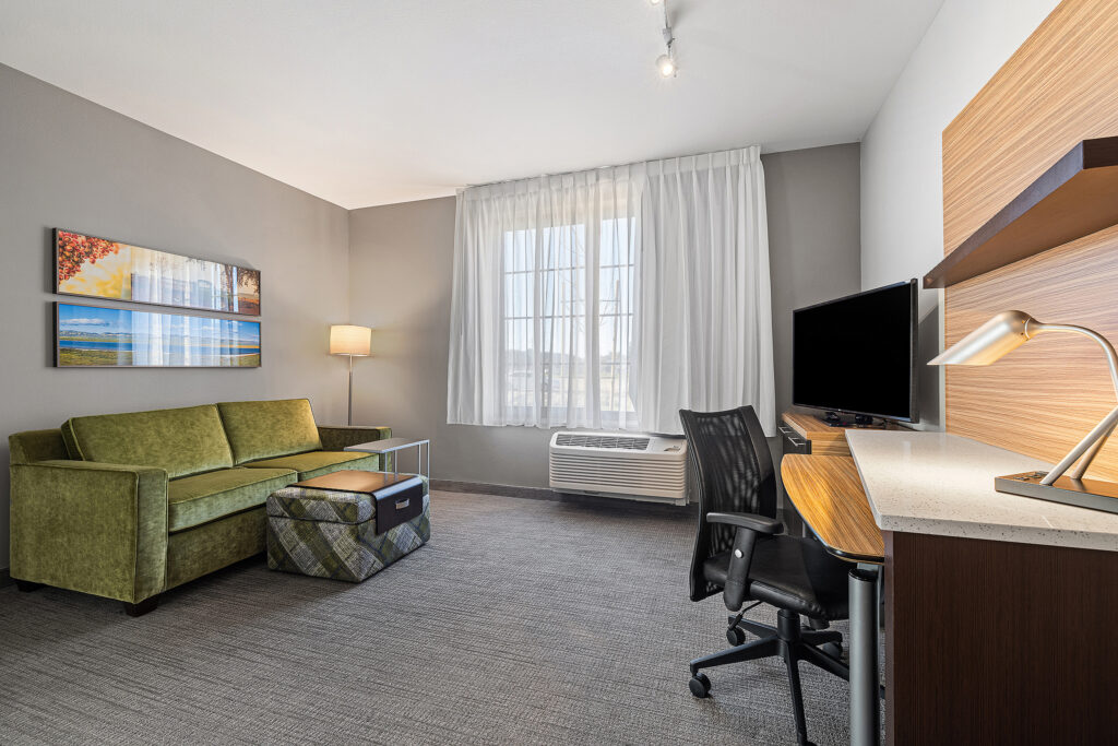 A modern Towneplace Suites hotel room in San Luis Obispo features a green sofa, ottoman, and tall lamp in the seating area. A work desk with an office chair, lamp, and TV is on the right. A large window with sheer curtains lets natural light brighten the gray-toned space elegantly.