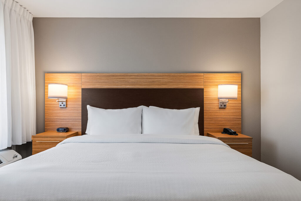 A neatly made bed with white linens is centered in a Towneplace Suites hotel room. The headboard features wooden accents and built-in lamps on each side. Two nightstands hold a phone and an alarm clock. The beige walls complement the open curtains, inviting the San Luis Obispo sunlight to stream in.