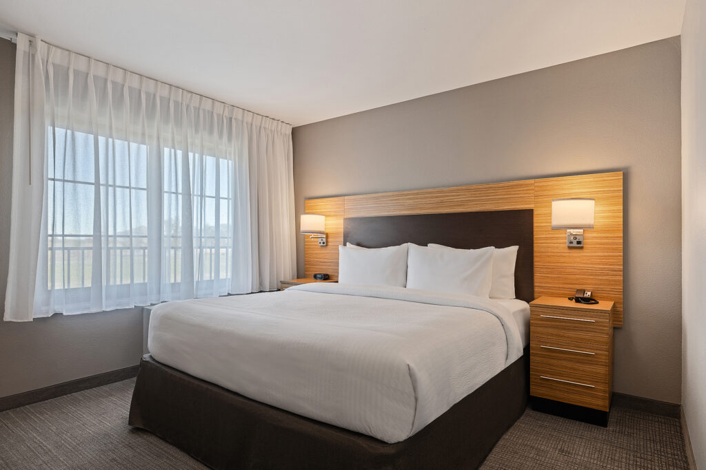 A modern hotel room at Towneplace Suites in San Luis Obispo features a large bed with white linens and a wooden headboard. Two matching nightstands with lamps flank the bed, while sheer white curtains on the window let natural light illuminate the calming, neutral-toned California ambiance.
