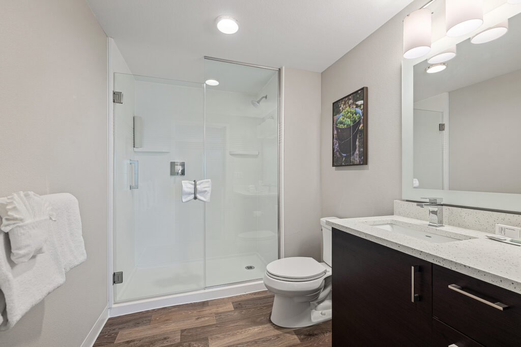 A modern bathroom at the Marriott in San Luis Obispo boasts a glass-enclosed shower with white tiles and a towel folded into a bow. A vanity with a light countertop and dark wood cabinets sits beside the toilet, while a framed picture adorns the light-colored wall.