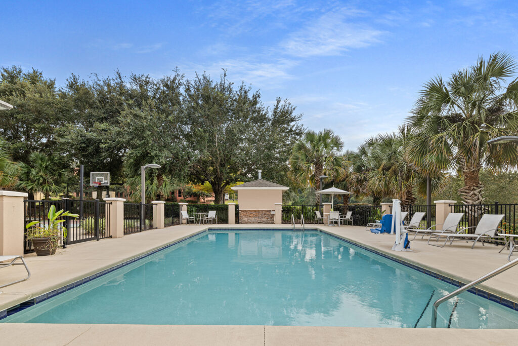 A rectangular outdoor swimming pool in Tallahassee, Florida, is surrounded by lounge chairs and trees. The pool is bordered by a black metal fence, with tall palm trees enhancing the backdrop. The clear sky has just a few clouds, creating a serene getaway perfect for an IHG hotel experience.
