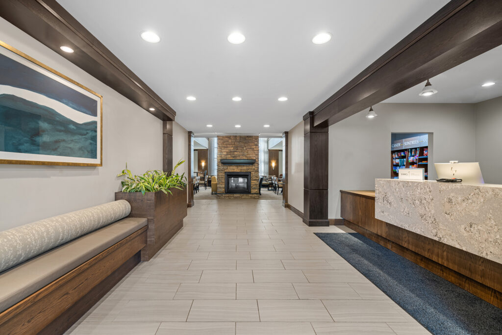 A modern Staybridge Suites hotel lobby in Tallahassee welcomes guests with its beige tiled floor and dark wood accents. You'll find a bench adorned with green plants on the left, a stone reception desk on the right, and a cozy seating area with a fireplace in the background.