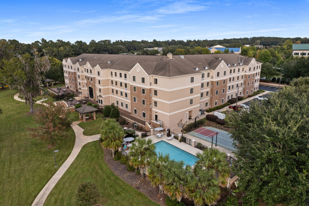 Aerial view of a large multi-story IHG residential building with brick and stucco exterior, reminiscent of Tallahassee's charm. Nestled among trees and landscaping, it boasts a swimming pool and tennis court in the foreground, with a clear blue sky overhead.