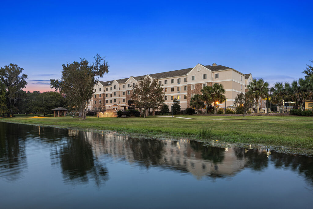 A modern, multi-story hotel with a beige and white exterior stands next to a calm, reflective body of water in Tallahassee. Surrounded by lush trees and a well-kept lawn, the Staybridge Suites are beautifully set under a clear blue sky during dusk.