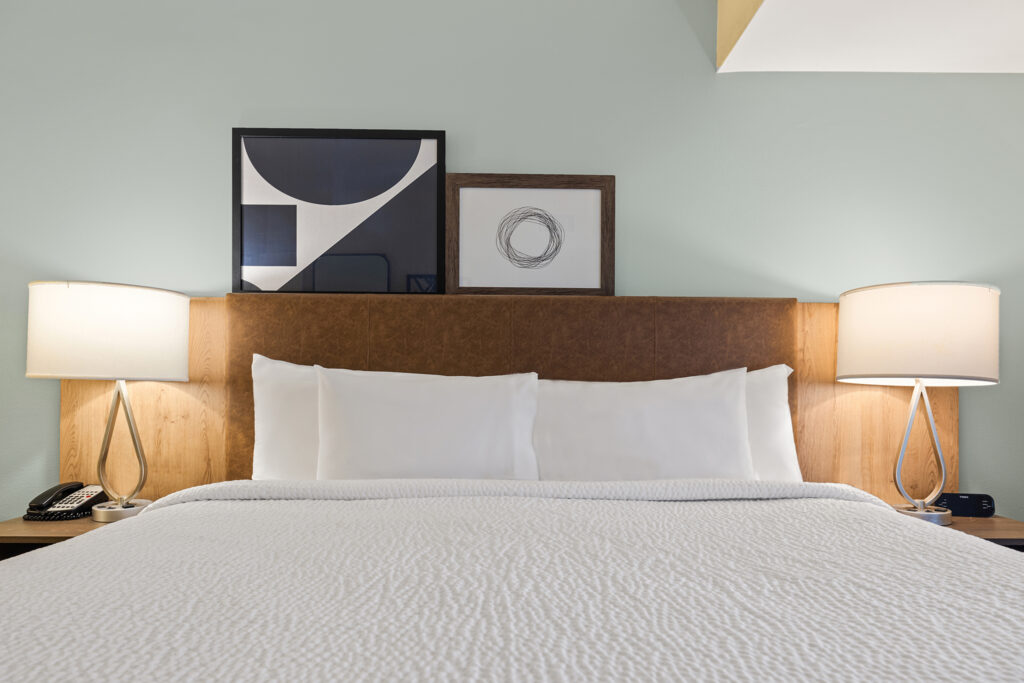 A neatly made bed with white linens is centered between two modern bedside lamps on wooden nightstands. Above the bed, three geometric art pieces hang on a light green wall, adding a minimalist touch to this Tallahassee hotel room.