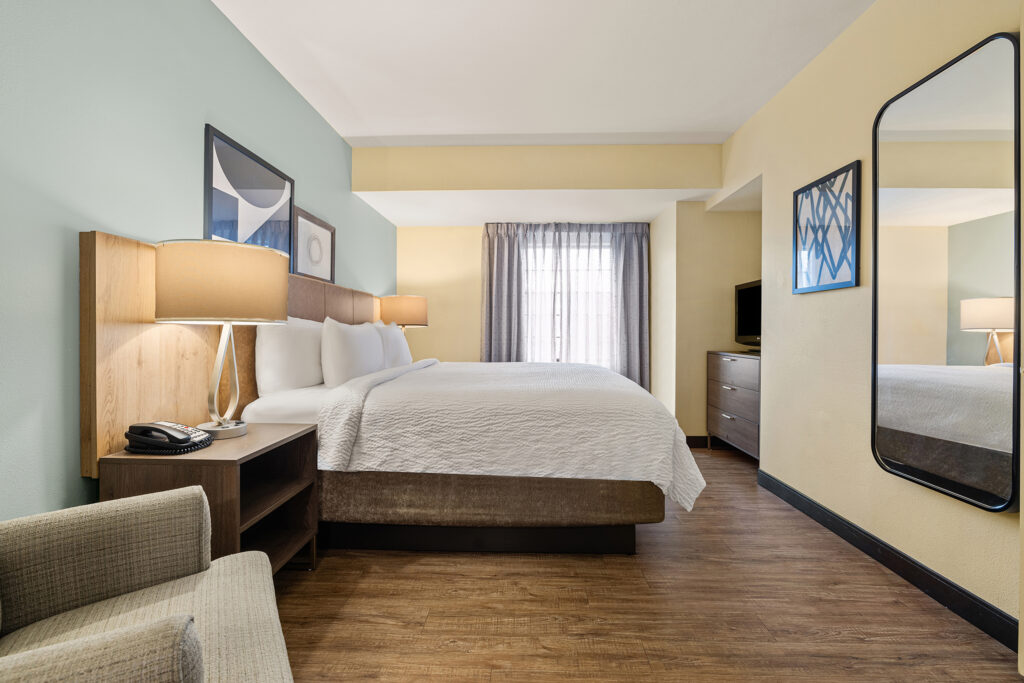 A modern Staybridge Suites hotel room welcomes you with a large bed adorned in white linens, flanked by wooden nightstands and lamps. Enjoy a soft armchair, wall-mounted mirror, dresser with TV, and window draped in sheer curtains. Your perfect Florida getaway awaits.