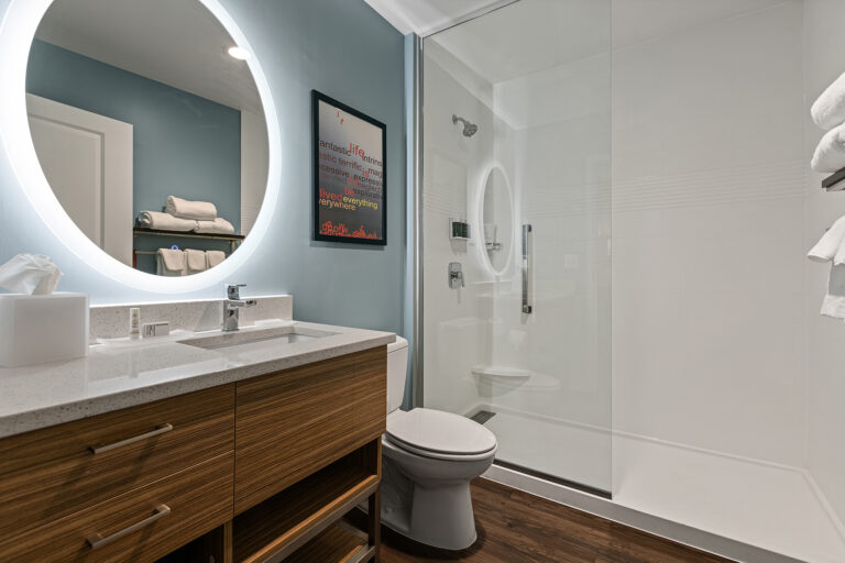 Experience a modern bathroom reminiscent of a luxury hotel, featuring a wooden vanity with a round, backlit mirror. A sleek glass-enclosed shower lies on the right, with neatly folded towels resting on open shelves. Blue-painted walls and a framed quote complete this Townplace Suites by Marriott-inspired oasis.