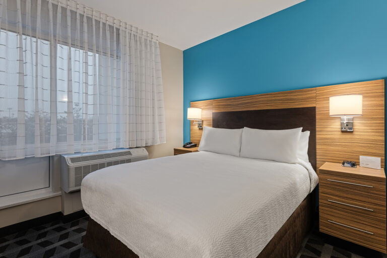 The TownPlace Suites hotel room features a large bed draped in white linens, flanked by wooden nightstands and lamps. A blue accent wall adds charm, while sheer curtains on the window flood the space with light. A convenient air conditioner is installed below for optimal comfort.