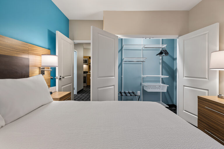 A Marriott hotel room features a neatly made bed with white linens, two bedside lamps on wooden tables, and an open closet with shelves and hangers. The walls are painted in light blue and beige tones.