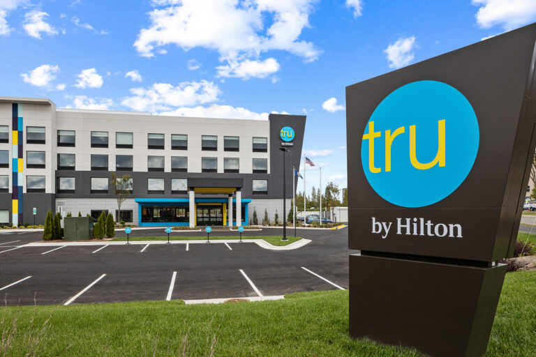 A modern hotel with the Tru by Hilton logo prominently displayed on a large sign captures attention in this striking piece of photography. The four-story building features a bright color scheme, multiple windows, and a welcoming entrance set against a partly cloudy sky.