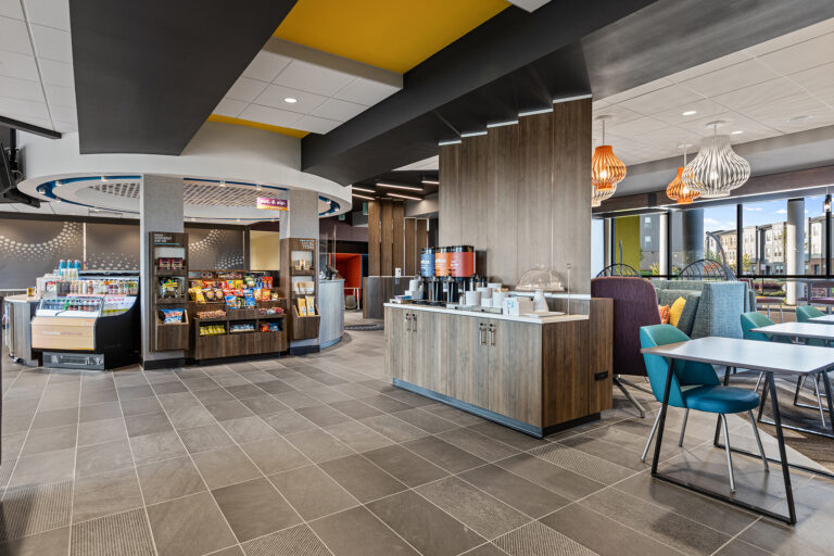 Bright and modern lounge area at Hilton, with a snack bar featuring various packaged items on shelves. A coffee station sits on a wooden counter, complete with cups and condiments. Contemporary chairs and tables are arranged beneath stylish hanging light fixtures, perfect for photography enthusiasts.