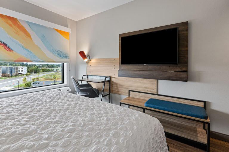 The Tru by Hilton offers a modern hotel room with a large bed, adorned in a textured white comforter. Opposite is a wall-mounted flat-screen TV and desk. A colorful window shade artfully frames the cityscape outside, perfect for capturing vibrant photography enthusiasts.