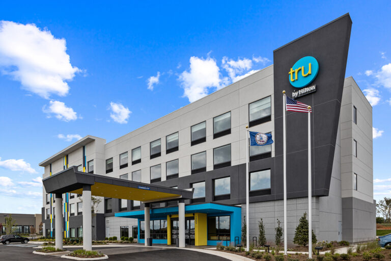 A modern Tru hotel with a geometric design showcases a striking blue and yellow entrance. Flags flutter in front against a clear sky dotted with clouds, perfect for photography. The four-story building is beautifully framed by landscaped greenery, inviting guests to relax.