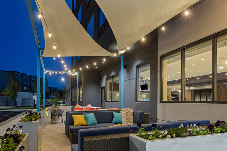 At dusk, this modern outdoor seating area resembles a Tru by Hilton patio, featuring a blue sectional sofa accented with orange and yellow cushions. Overhead string lights enhance the cozy ambiance, while sail shades and potted plants provide coverage amidst the scenic cityscape.
