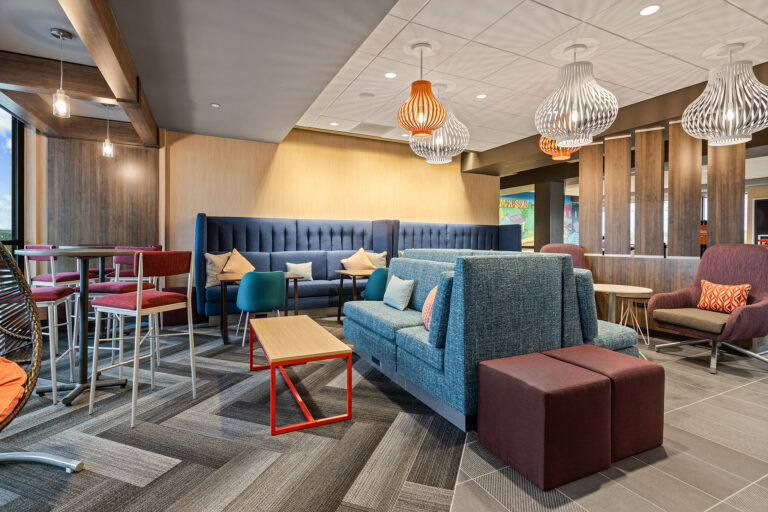 This contemporary lounge, reminiscent of a stylish Hilton hotel, features modern furniture with blue sofas, padded chairs, colorful cushions, and a mix of pendant lights. Geometric patterns on the carpet complement wooden partitions and tables, creating a cozy yet vibrant atmosphere perfect for photography.