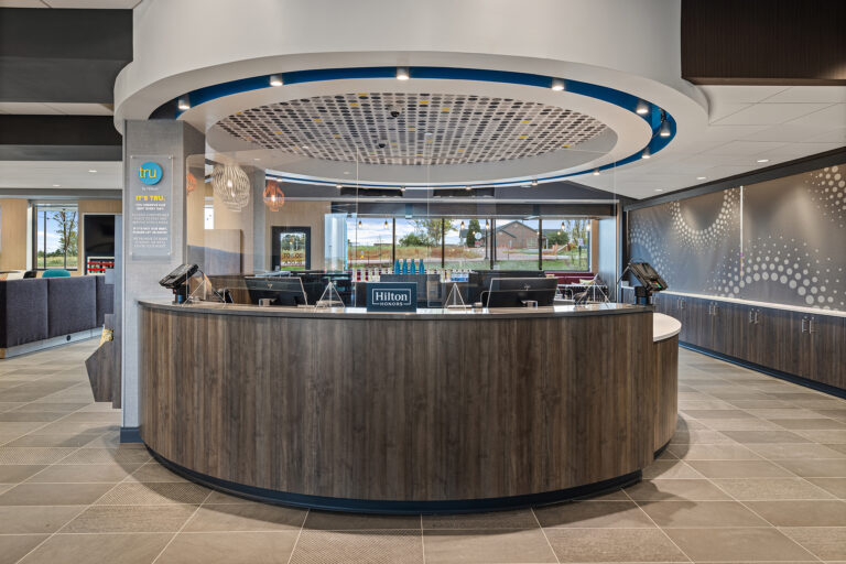 Hotel lobby with a modern, circular front desk featuring a wood finish and multiple check-in stations. The ceiling has a unique circular design. Large windows in the background provide natural light, and there is a seating area on one side.