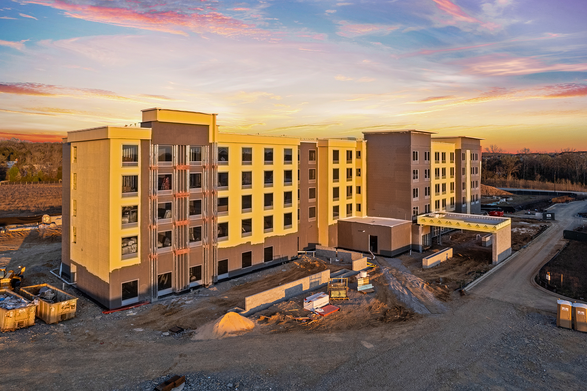 An aerial view captures the construction site of a multi-story building at sunset, with the silhouette reminiscent of a future Hilton. Incomplete facades and scattered materials rest under a sky awash in vibrant orange and pink, contrasting beautifully with the earthy tones below.