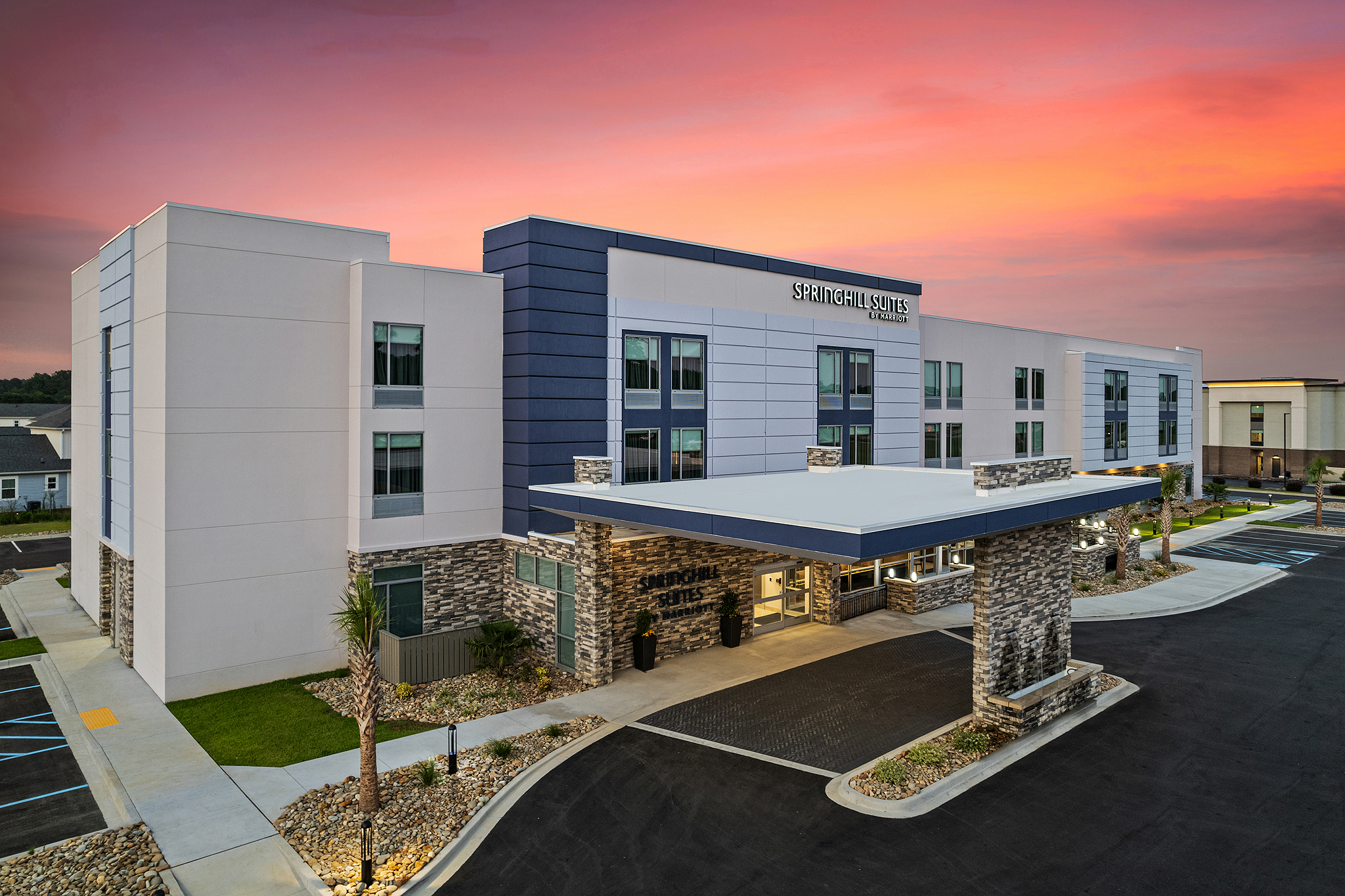 A modern hotel building with stone and blue accents is shown at dusk under a vibrant pink and orange sky in Camden, South Carolina. The entrance features a covered drive, landscaped areas, and well-lit windows that embody true Southern hospitality.