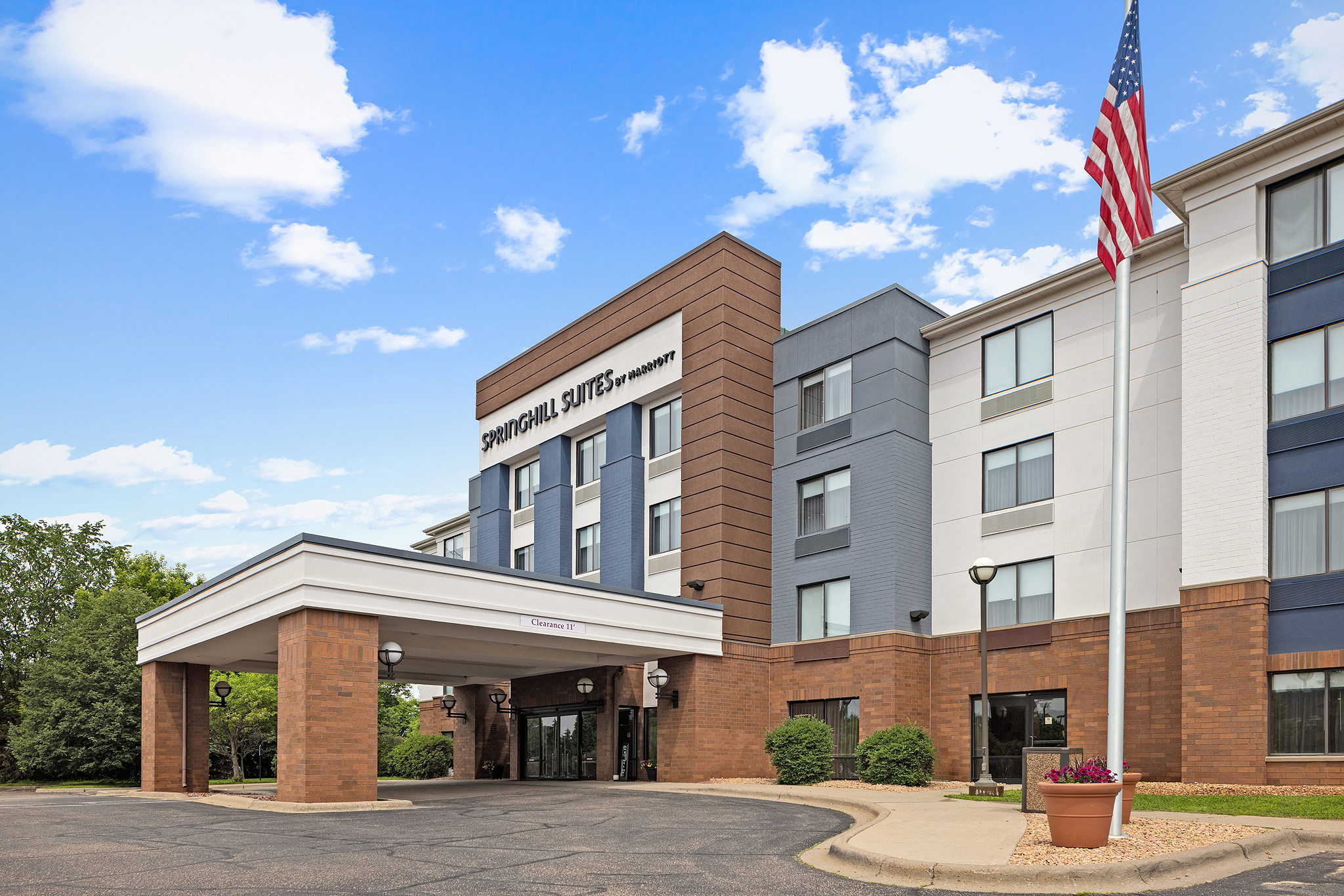 A SpringHill Suites by Marriott hotel in Minneapolis, MN with a modern exterior featuring brick and white paneling, a covered entrance, and an American flag on a pole near the entrance under a partly cloudy sky.