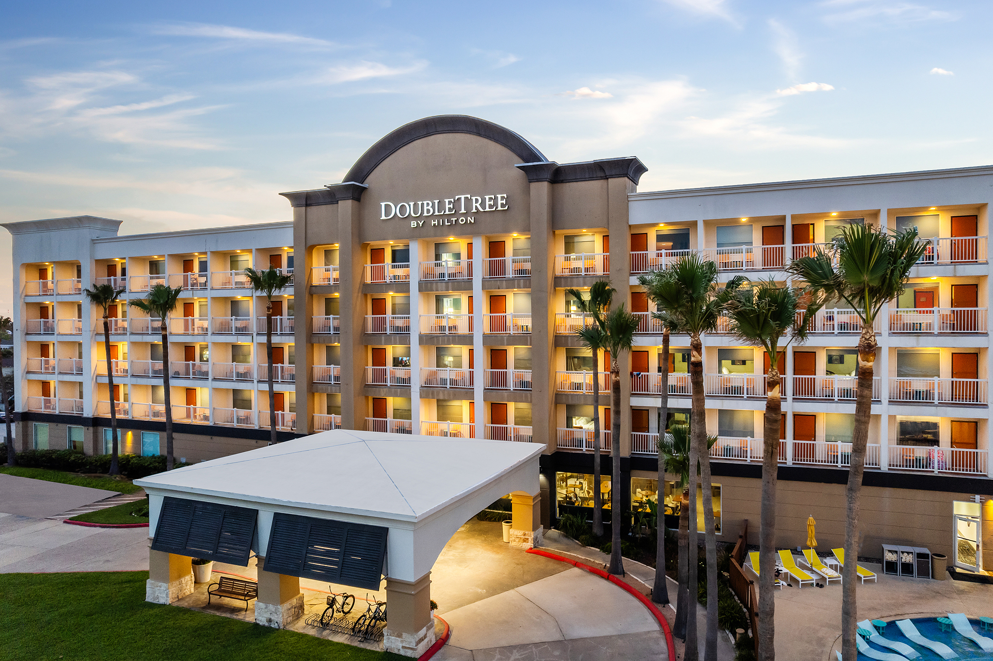 A four-story Hilton hotel is shown at sunset, with illuminated balconies, palm trees in front, and a covered entrance. The partly cloudy sky adds to the ambiance, while a glimpse of the swimming pool area hints at relaxing beachside stays.