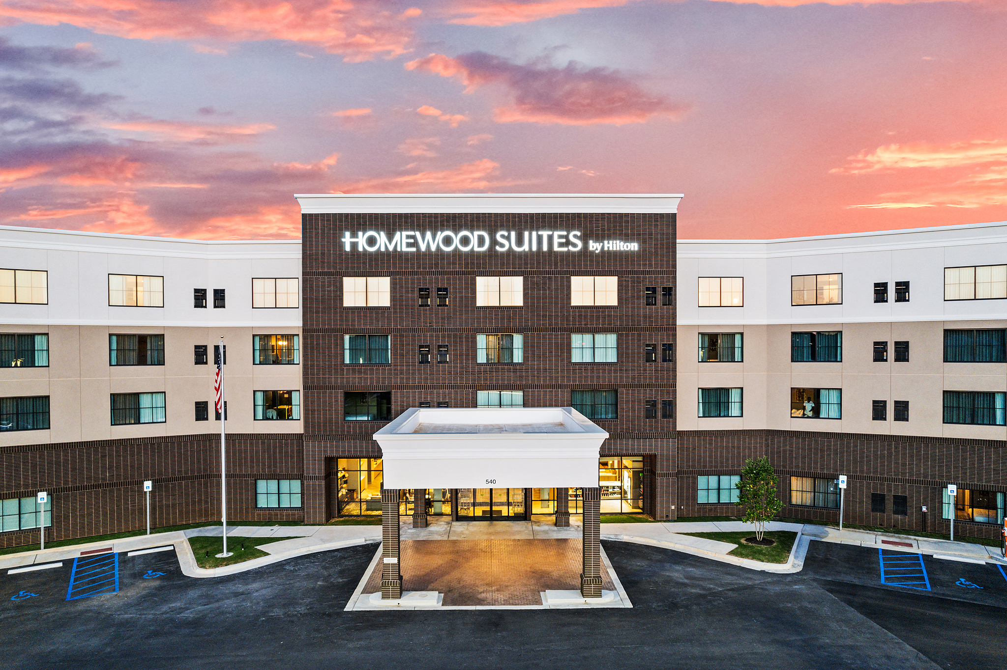 A modern Homewood Suites by Hilton hotel with a curved facade, large windows, and a covered entrance welcomes guests with signature Hilton hospitality. The vibrant sunset frames accessible parking spaces in the foreground.