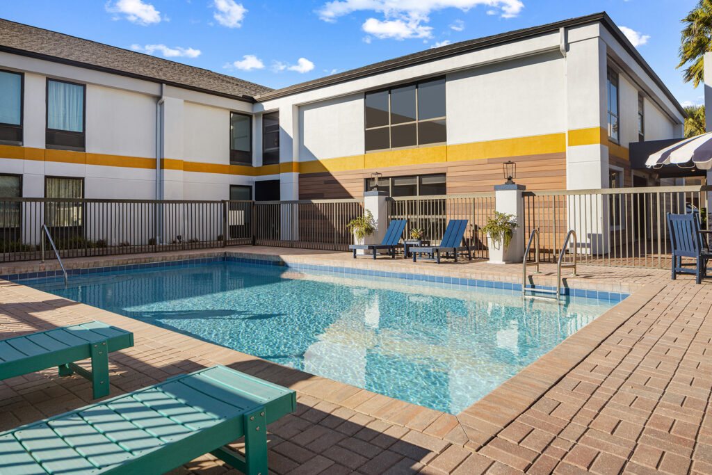 Outdoor swimming pool at the City Express hotel in Yulee, surrounded by a brick patio, green lounge chairs, and a metal fence, with a modern two-story building in the background under a blue sky with scattered clouds.