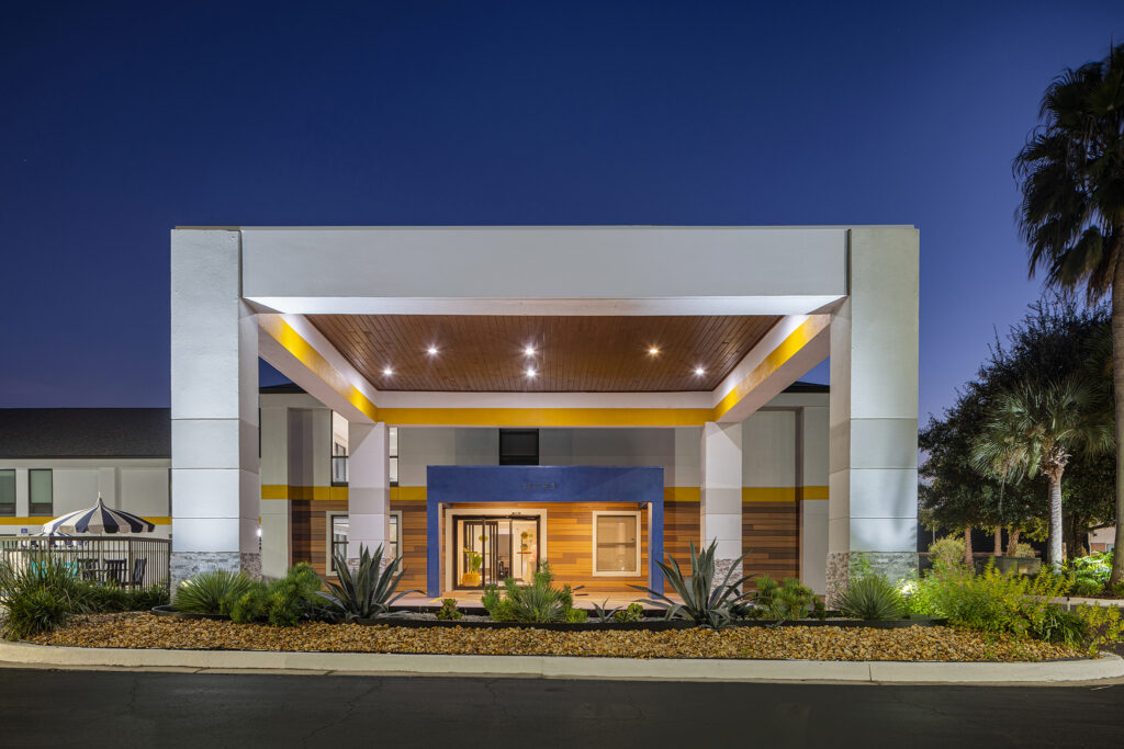 A modern Marriott hotel entrance in Yulee, Florida, shines at night with a large covered driveway, geometric design, landscaped plants, white walls, yellow and blue accents, and inviting glass doors.