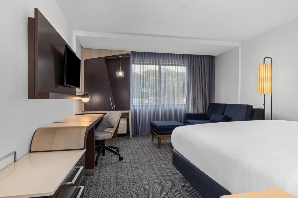 Modern Marriott hotel room with a neatly made bed, desk and chair, wall-mounted TV, blue sofa and ottoman, floor lamp, and large window with sheer curtains filling the space with natural light—perfect for hotel photography.