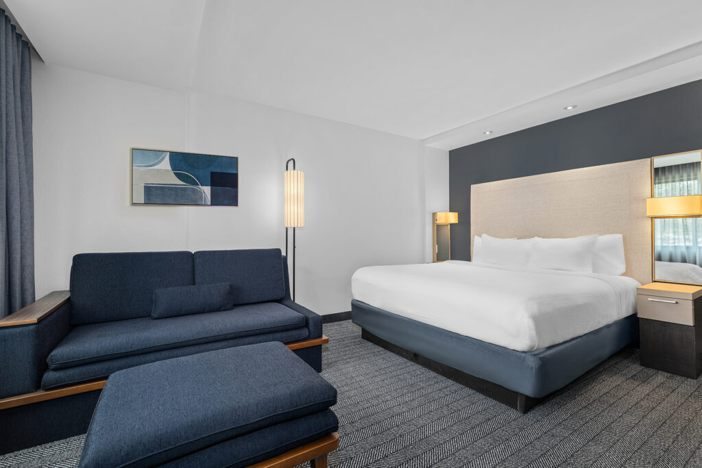 A modern hotel room in Oxford, MS features a large bed with white linens, a blue sofa, ottoman, two nightstands with lamps, floor lamp, and abstract wall art—perfect for hospitality photography with its minimalist and clean decor.