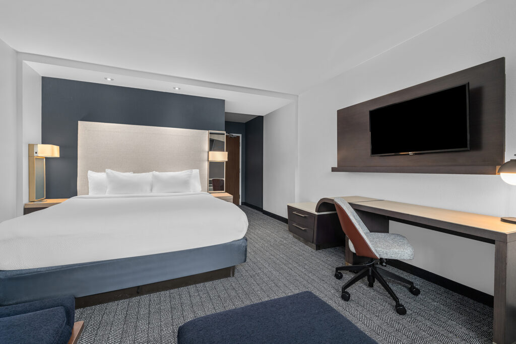 Modern Marriott hotel room with a neatly made bed, a large headboard, wall-mounted TV, desk and chair, bedside lamps, and neutral-colored decor. The spacious room overlooks the courtyard and is ideal for hotel photography.