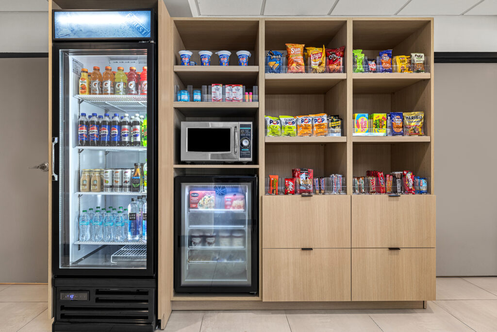 A snack area in a modern, light-wood cabinet at a Yulee hotel, featuring a fridge of drinks, microwave, small fridge with food, and shelves stocked with chips, snacks, and candy—perfect for your Florida getaway.