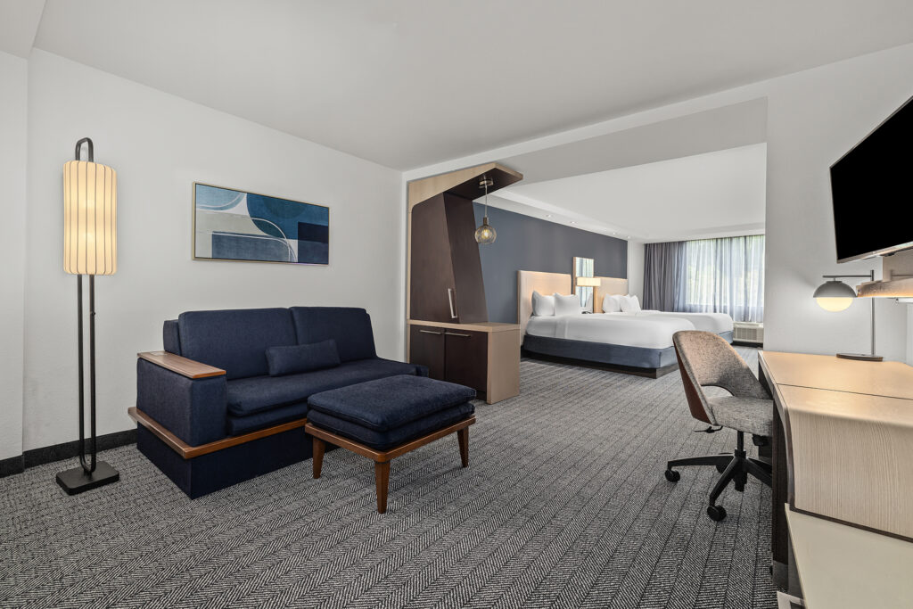 Modern hotel room in Oxford, MS, with a blue sofa and ottoman, desk, floor lamp, abstract wall art, and bed near large windows. Perfect for Marriott hotel photography, the contemporary decor features neutral tones throughout.