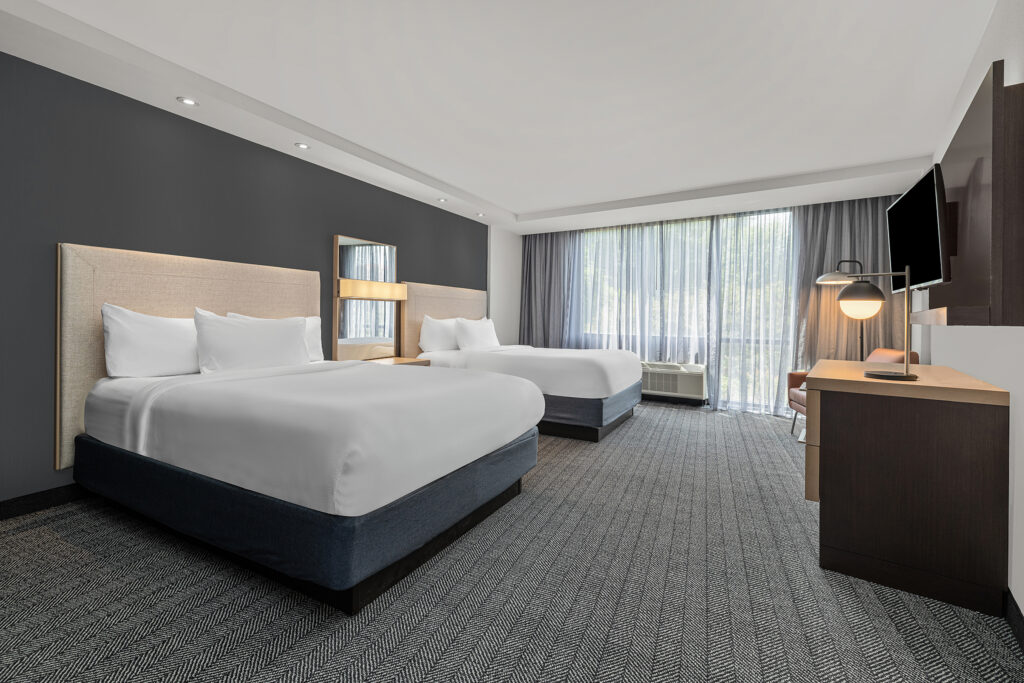 Modern hotel room at Marriott in Oxford, MS featuring two queen beds, a large window with sheer curtains, a flat-screen TV, desk, and seating area. Captured with hospitality photography flair, the minimalist decor showcases neutral tones and carpeted flooring.
